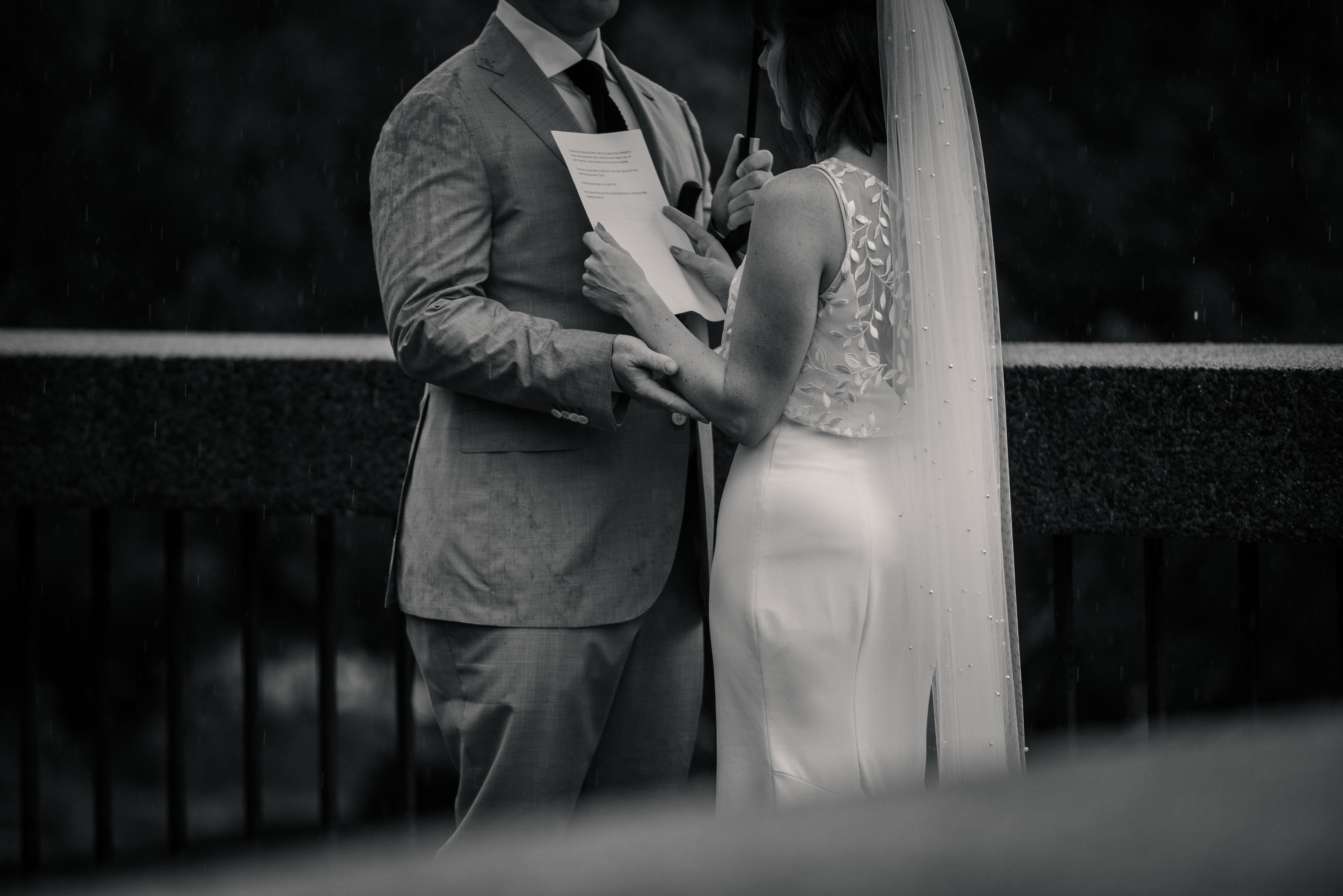 A groom holds his bride tight while she reads her vows to him under an umbrella in the rain