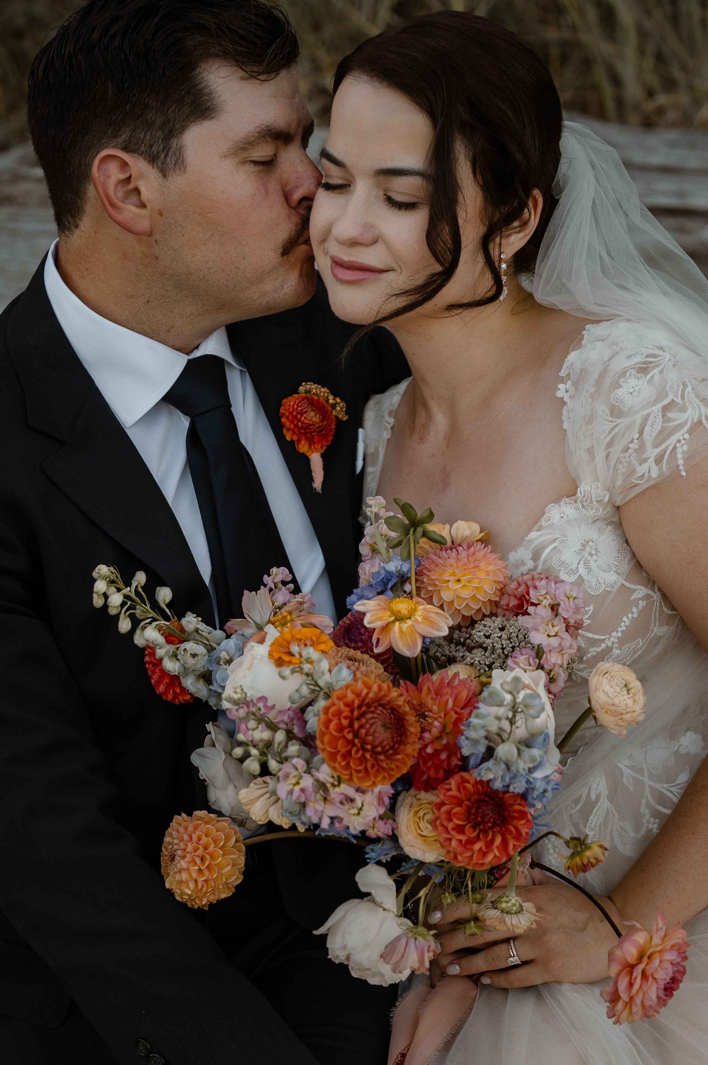 Groom kisses his bride on the cheek while she has her eyes closed and holds a beautiful bouquet of flowers