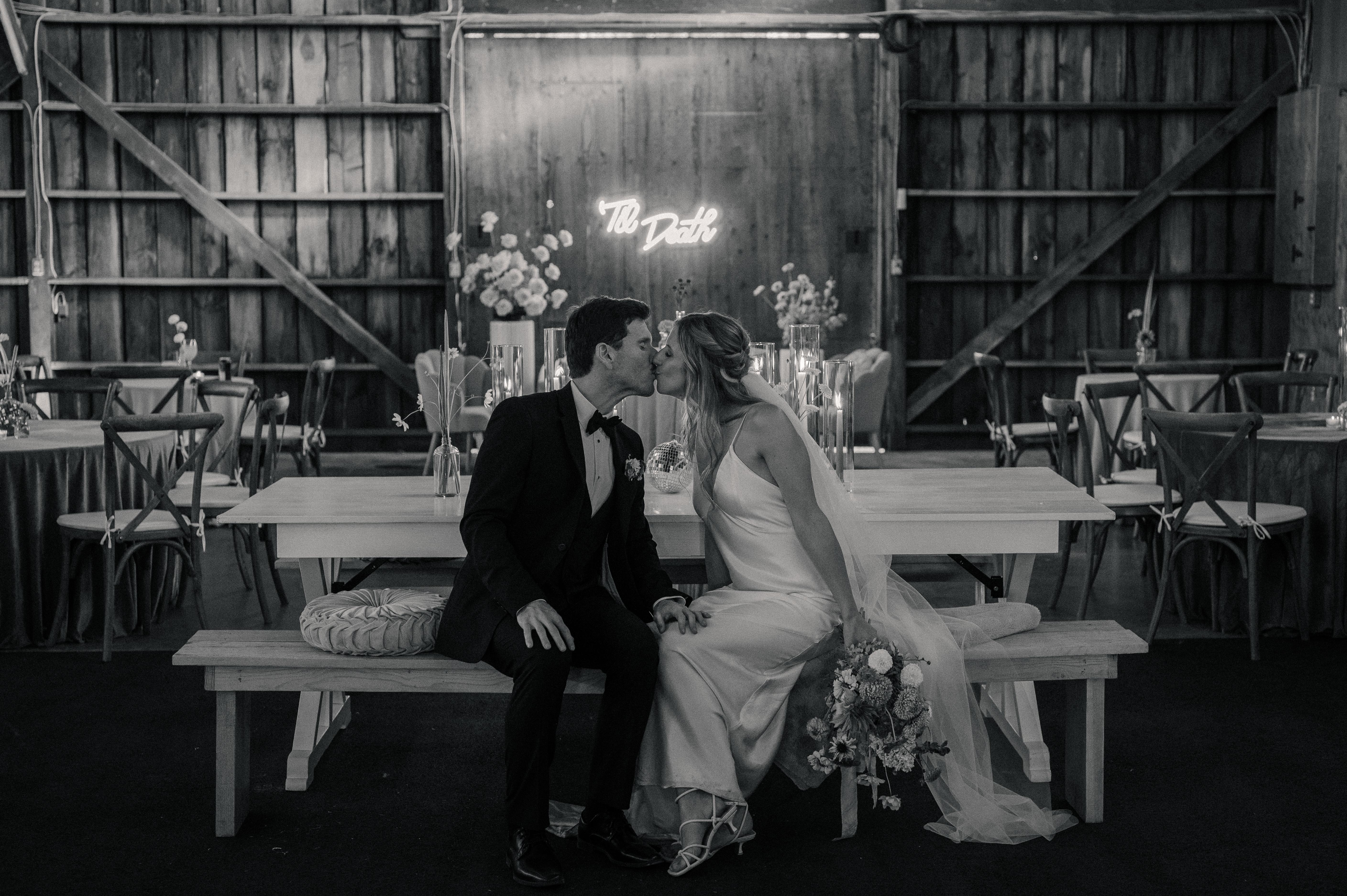 Couple sits together in their reception space in front of a sign that reads Till Death