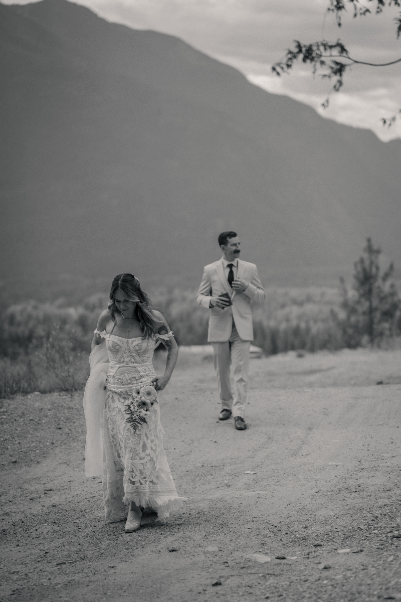 Bride and groom walkings a dirt path in the mountains, while the tuck sneakly tucks a flask back into his jacket pocket in Pemberton BC